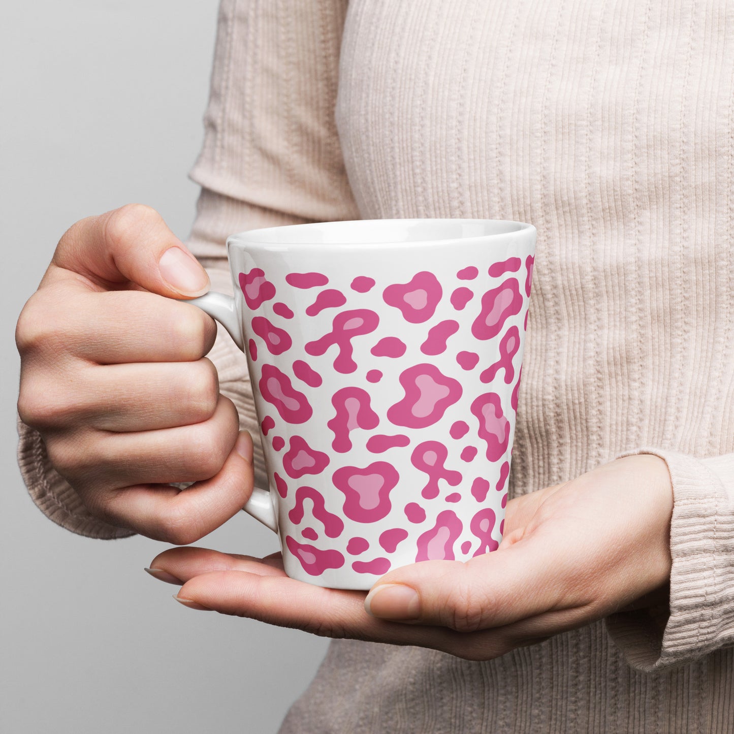 A person in a beige ribbed sweater holds a white ceramic mug with a pink leopard print pattern against a plain light gray background.