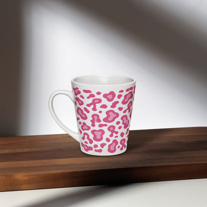 A white ceramic latte mug with a pink leopard print pattern, resting on a dark wooden board against a softly lit gray background.