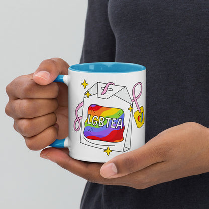 Hands holding a white ceramic LGBTea mug with a blue handle and interior, featuring a rainbow tea bag illustration with pink ribbons and sparkles.
