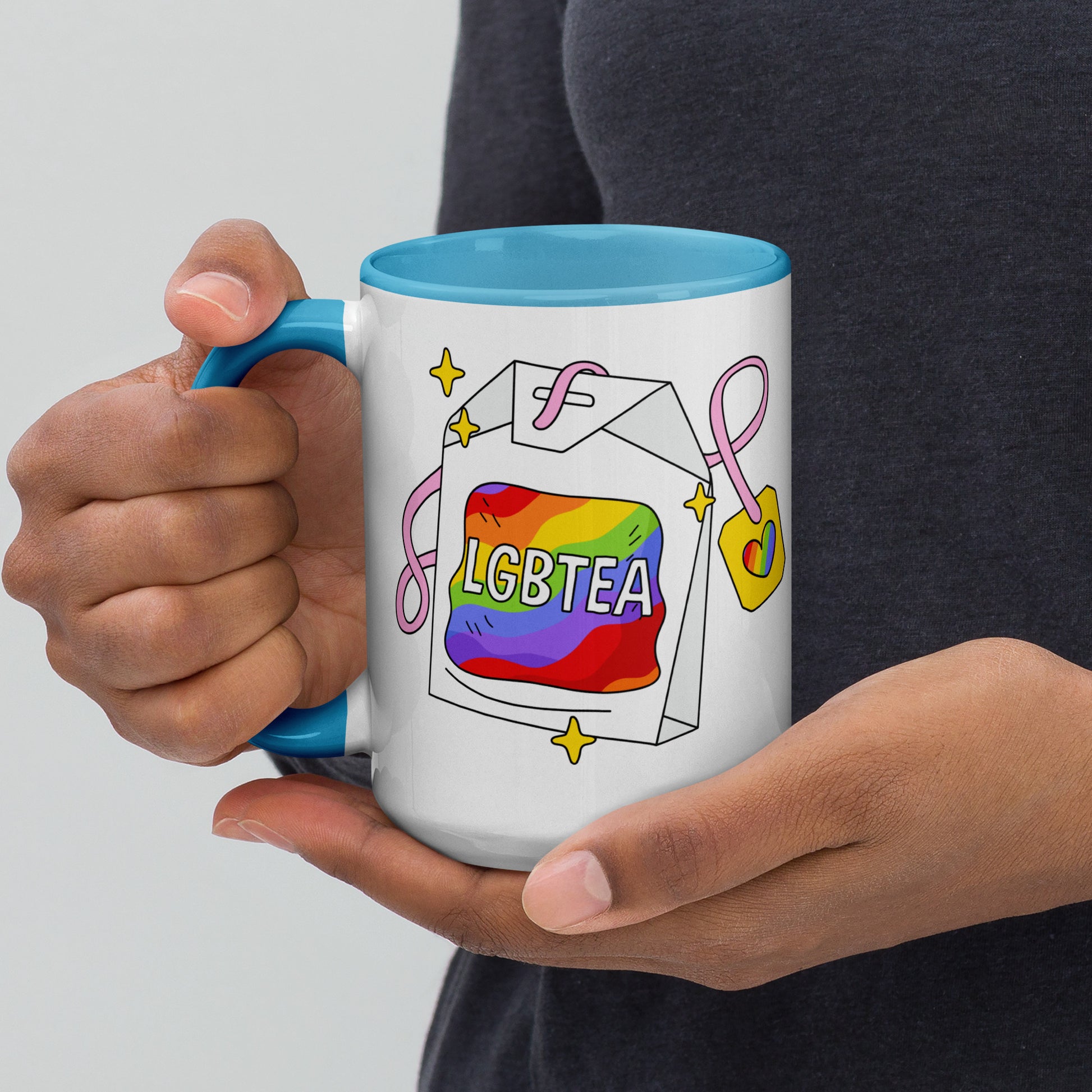 Hands holding a white ceramic LGBTea mug with a blue handle and interior, featuring a rainbow tea bag illustration with pink ribbons and sparkles.