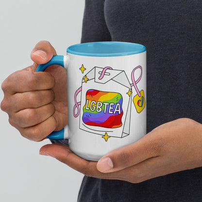Hands holding a white ceramic LGBTea mug with a blue handle and interior, featuring a rainbow tea bag illustration with pink ribbons and sparkles.