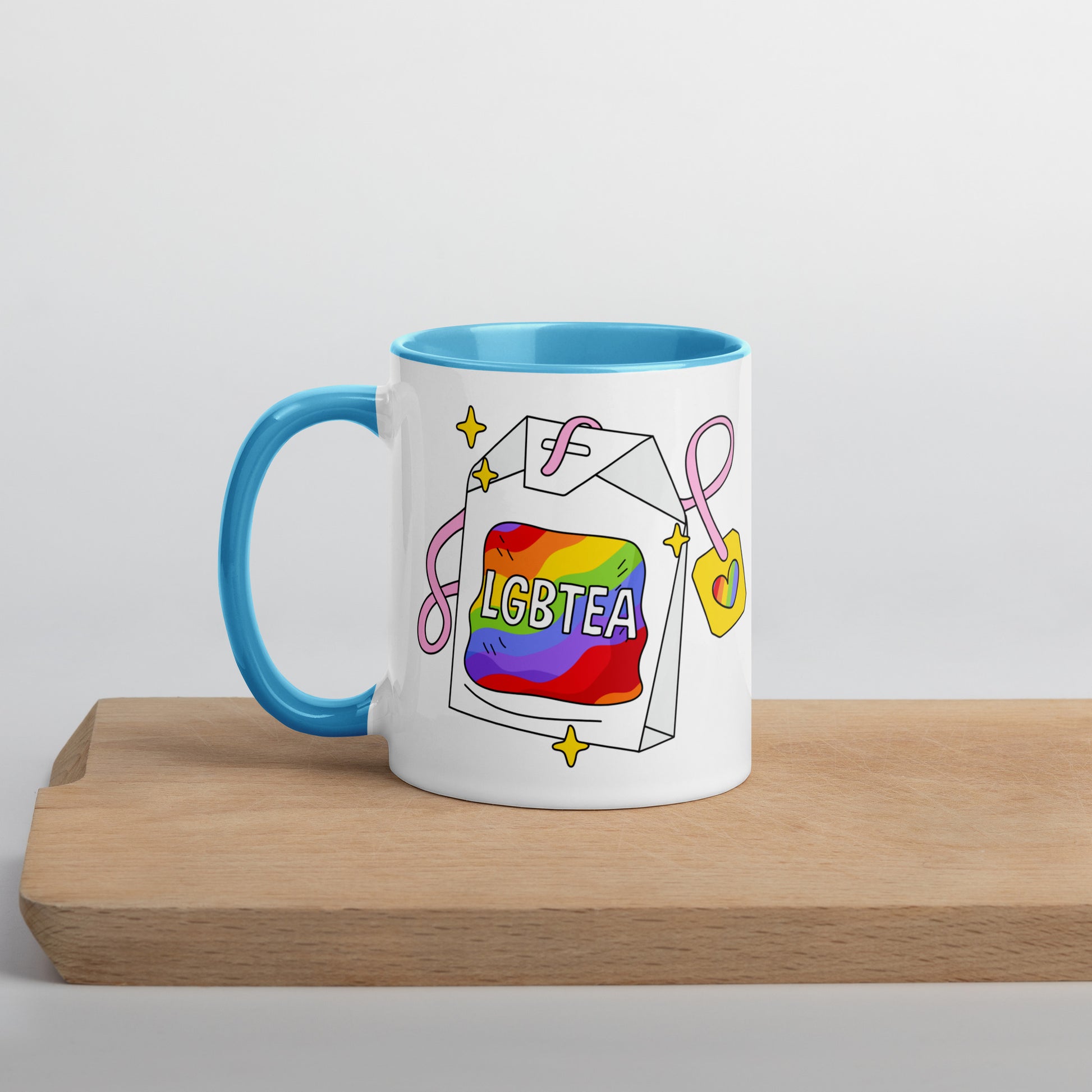White ceramic 11oz LGBTea mug with blue handle and interior, featuring a rainbow tea bag illustration with pink ribbons and sparkles.
