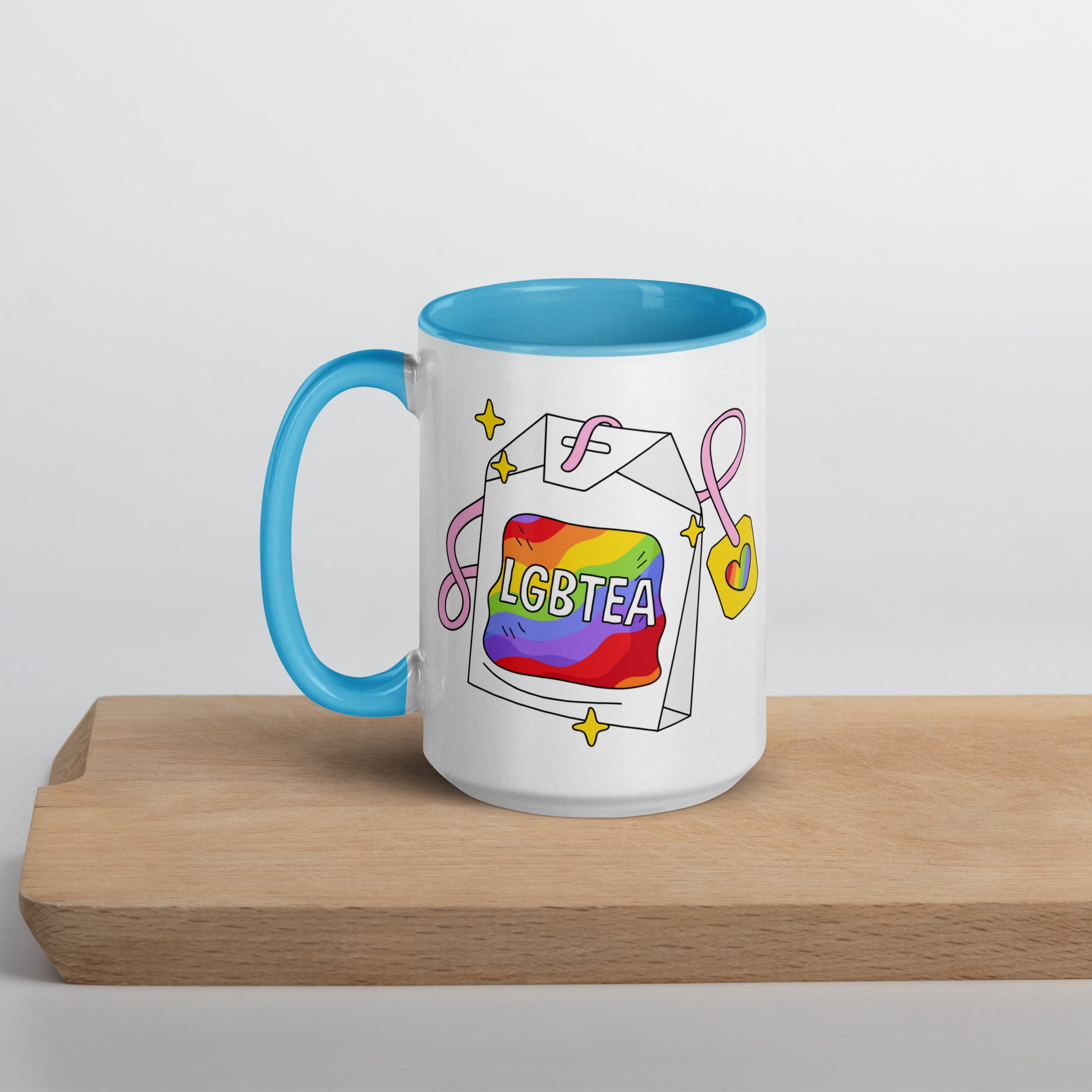 White ceramic 15oz LGBTea mug with blue handle and interior, featuring a rainbow tea bag illustration with pink ribbons and sparkles.