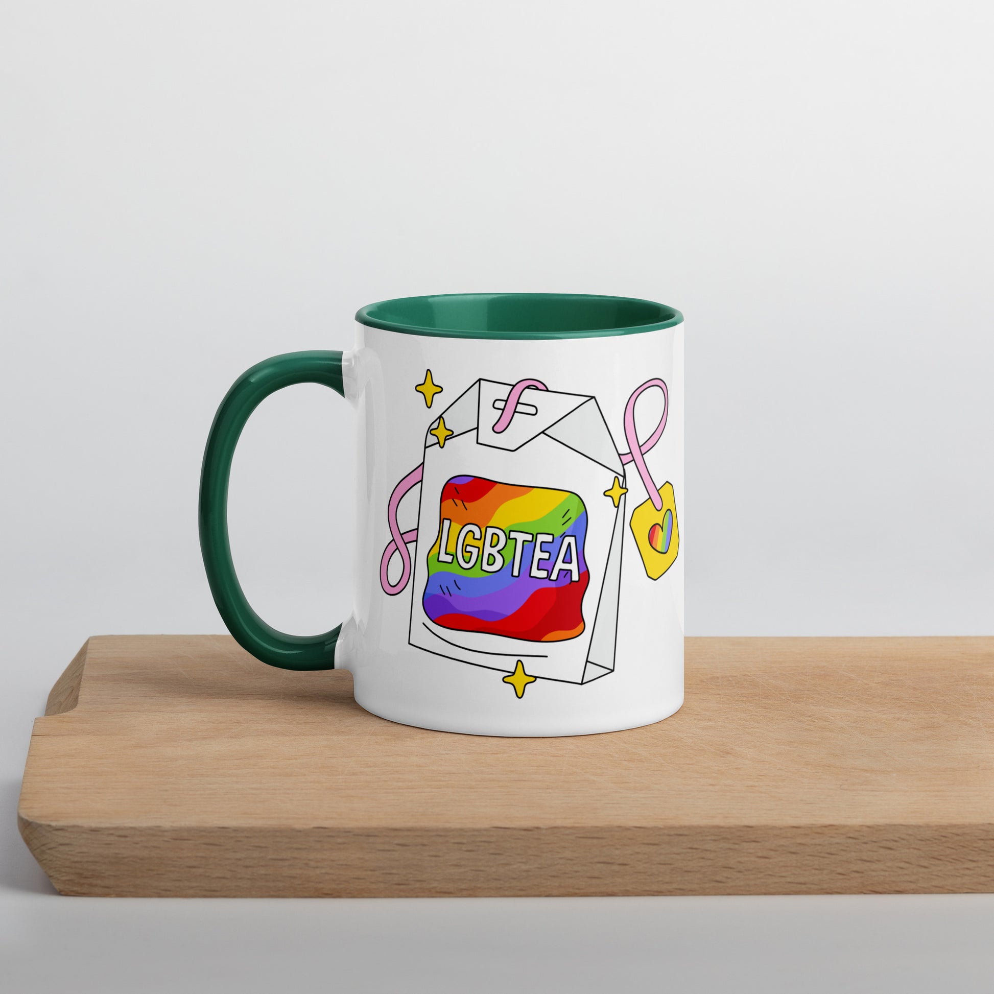 White ceramic 11oz LGBTea mug with dark green handle and interior, featuring a rainbow tea bag illustration with pink ribbons and sparkles.
