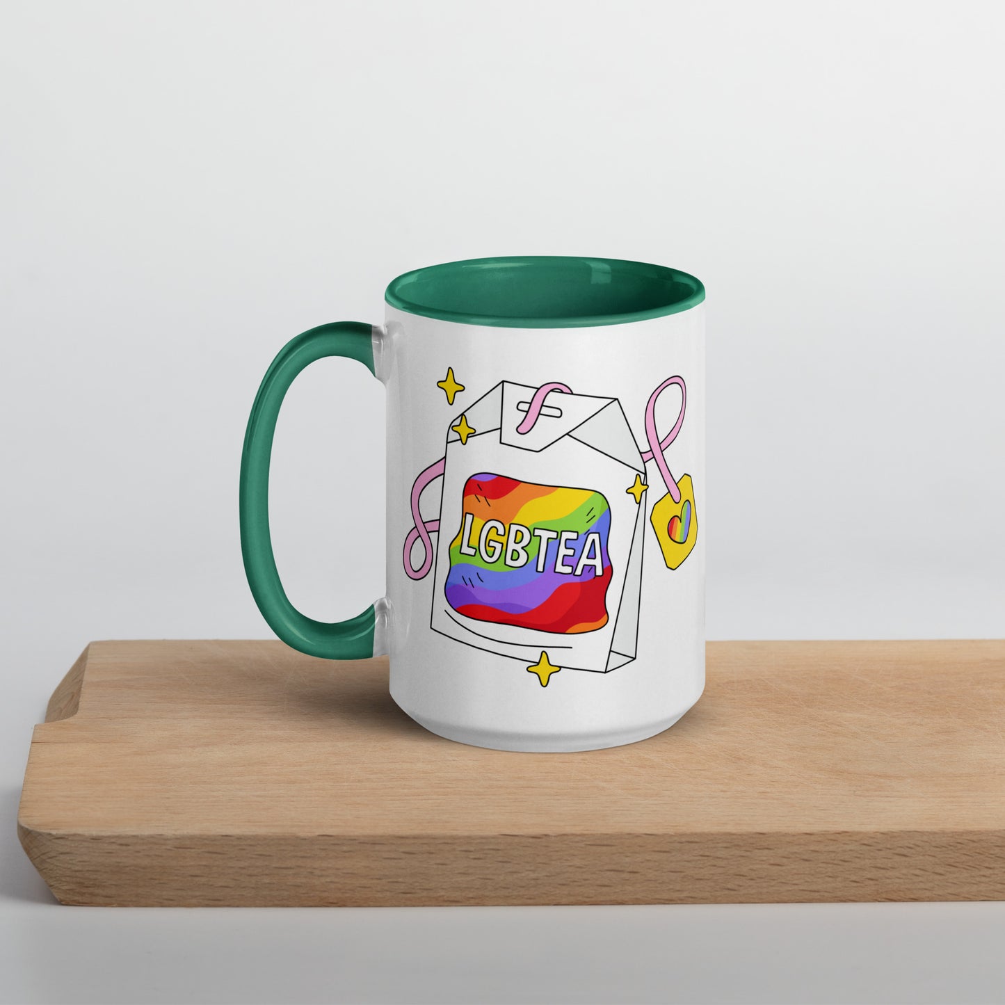 White ceramic 15oz LGBTea mug with dark green handle and interior, featuring a rainbow tea bag illustration with pink ribbons and sparkles.