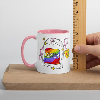 Pink-handled white ceramic LGBTea mug on a wood board beside a vertical ruler, shown at about 6 inches tall to display its size. The rainbow tea bag design with pink ribbons and sparkles faces forward.