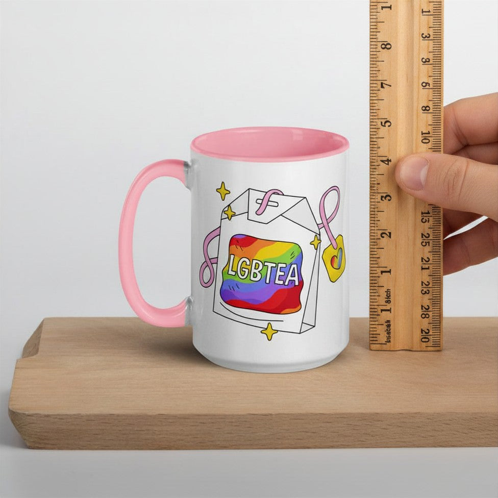White ceramic LGBTea mug with pink interior and handle, measured against an upright ruler on a wooden board to show its height. Rainbow tea bag illustration with pink accents and a small heart charm.