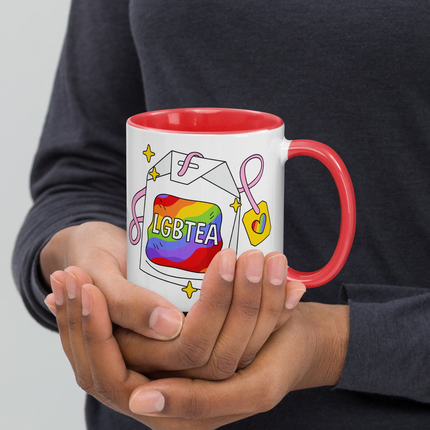 Hands holding a white ceramic LGBTea mug with a red handle and interior, featuring a rainbow tea bag illustration with pink ribbons and sparkles.