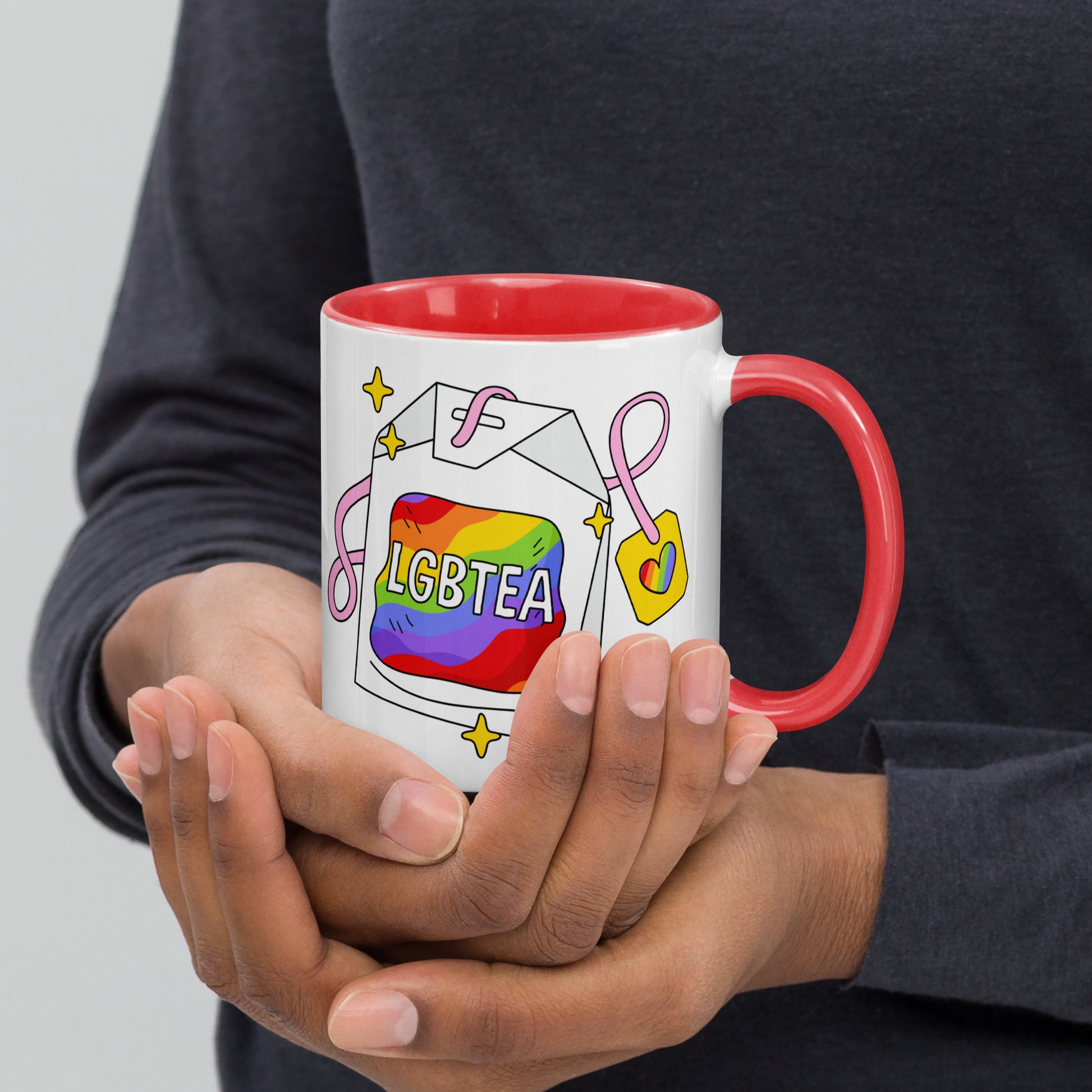 Hands holding a white ceramic LGBTea mug with a red handle and interior, featuring a rainbow tea bag illustration with pink ribbons and sparkles.