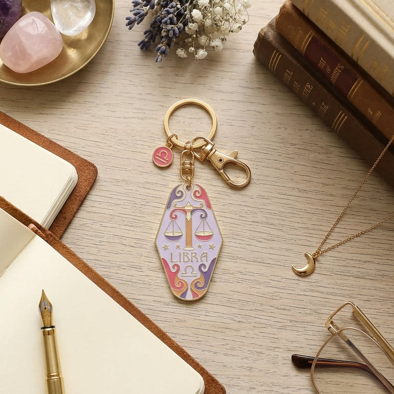 A Libra-themed keychain with a colorful scale-of-judgment pendant, gold-tone clasp, and small pink charm, displayed on a light wood desk with books, crystals, glasses, and a notebook.