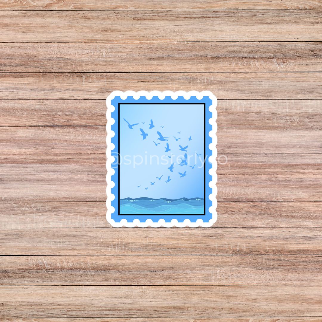 A rectangular die-cut sticker with a scalloped white border, featuring a blue sky with flying birds and a blue horizon, placed on a light brown wood plank background.