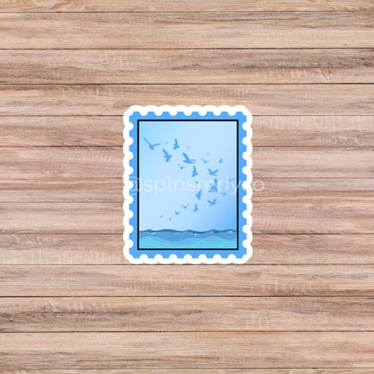 A rectangular die-cut sticker with a scalloped white border, featuring a blue sky with flying birds and a blue horizon, placed on a light brown wood plank background.