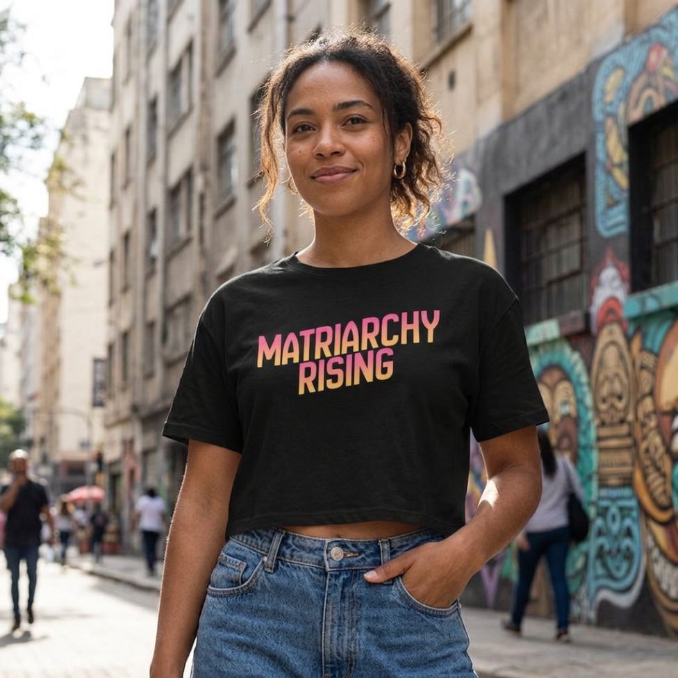 Woman wearing a black Matriarchy Rising crop top with sunrise-gradient lettering, paired with high-waisted jeans on a city street.