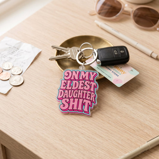 A pink and purple acrylic keychain with the text 'ON MY ELDEST DAUGHTER SHIT' in white and purple lettering, attached to a key ring with keys and a car key fob, displayed on a light wood desk surrounded by lifestyle accessories.