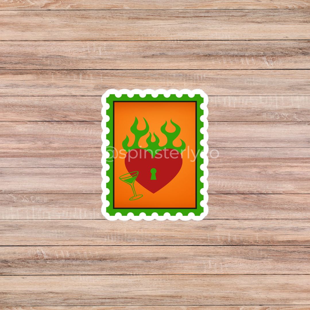 A rectangular die-cut sticker with a green scalloped border, featuring a red heart with a keyhole and green flames on an orange background, placed on a light brown wood plank surface.