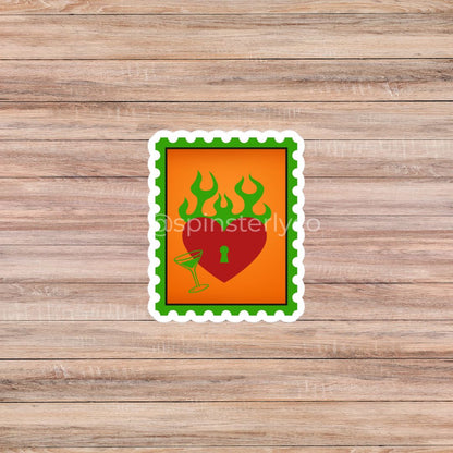 A rectangular die-cut sticker with a green scalloped border, featuring a red heart with a keyhole and green flames on an orange background, placed on a light brown wood plank surface.