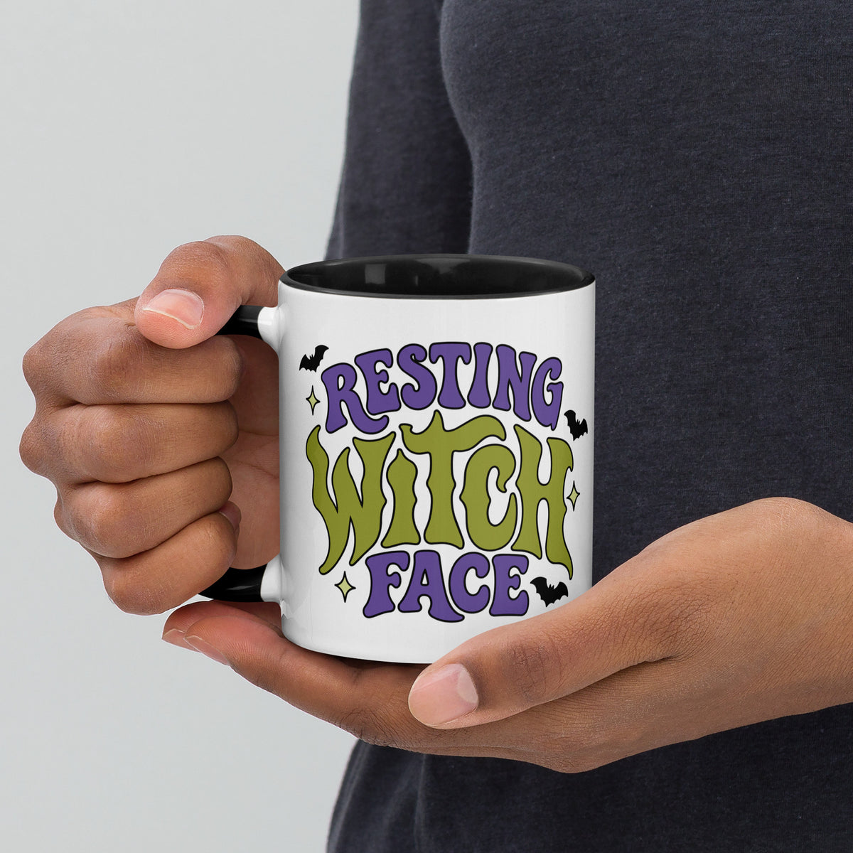 A person holding a white ceramic mug with a black handle and interior. The mug features a colorful graphic that reads 'RESTING WITCH FACE' in purple and green lettering with small black bats and star accents.