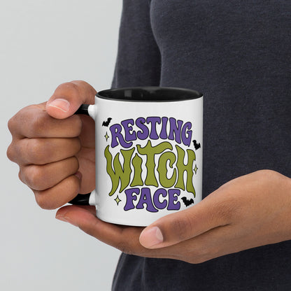 A person holding a white ceramic mug with a black handle and interior. The mug features a colorful graphic that reads 'RESTING WITCH FACE' in purple and green lettering with small black bats and star accents.