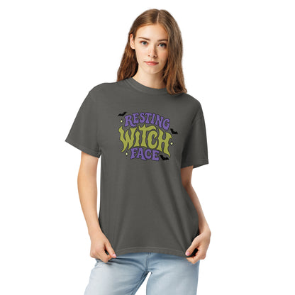 A woman modeling a dark gray t-shirt with a colorful graphic that reads 'RESTING WITCH FACE' in purple and lime green, paired with light blue jeans, against a plain white background.