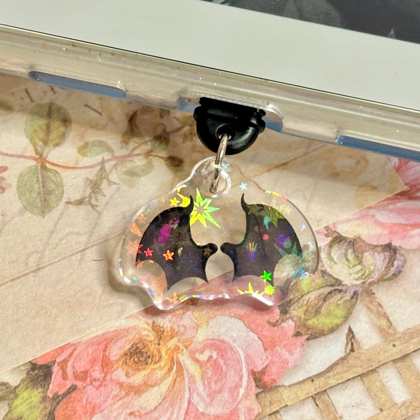 Decorative phone charm with star and batwing design on a floral-patterned surface