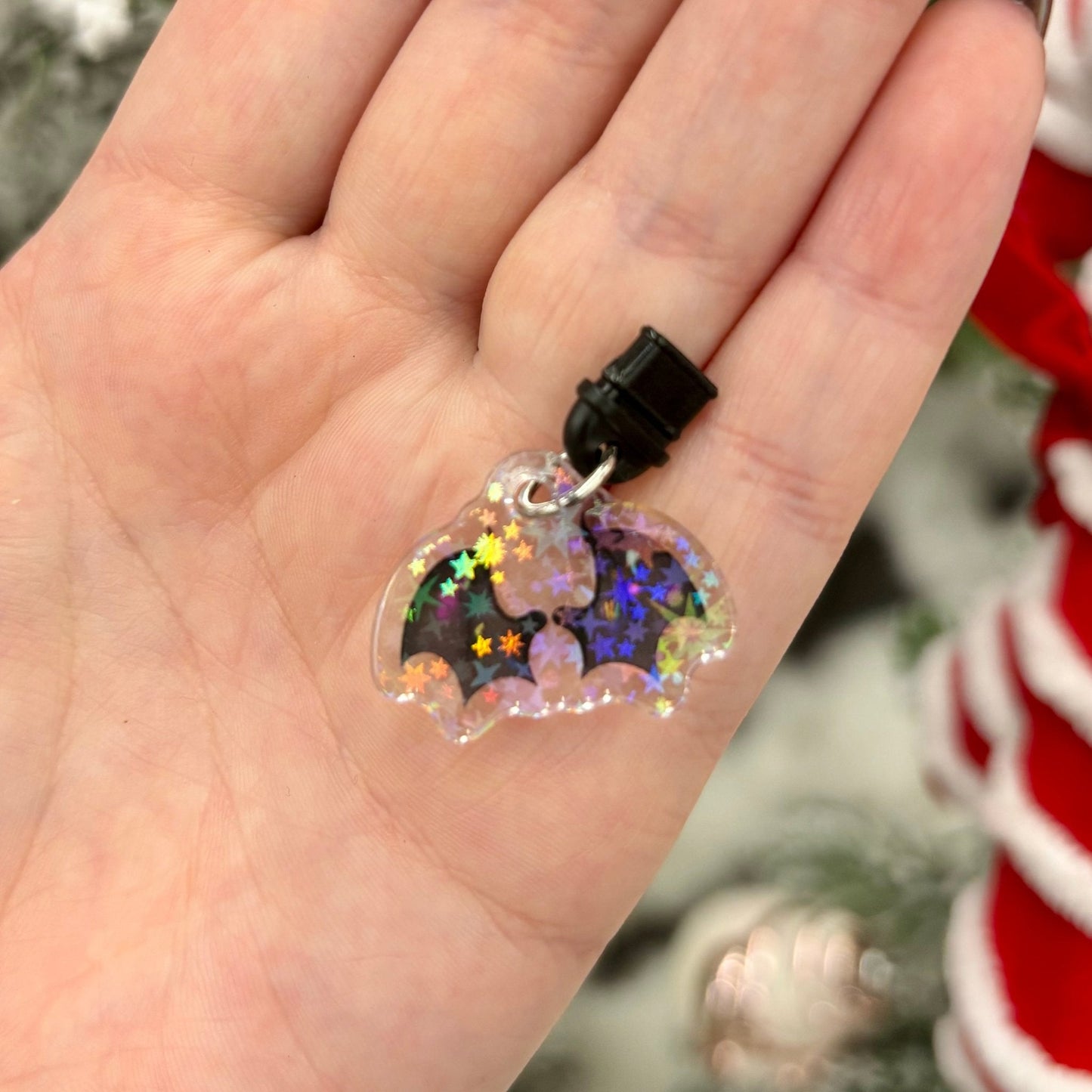 Hand holding a small, clear plastic phone charm with wings and star designs against a blurred Christmas tree background.