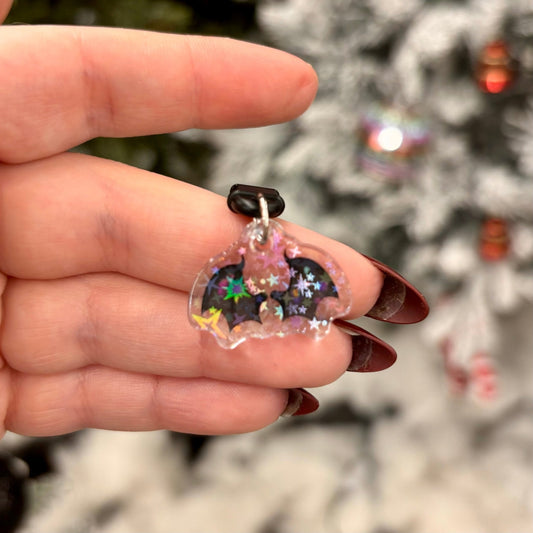 Hand holding a small, clear plastic phone charm with wings and star designs against a blurred Christmas tree background.