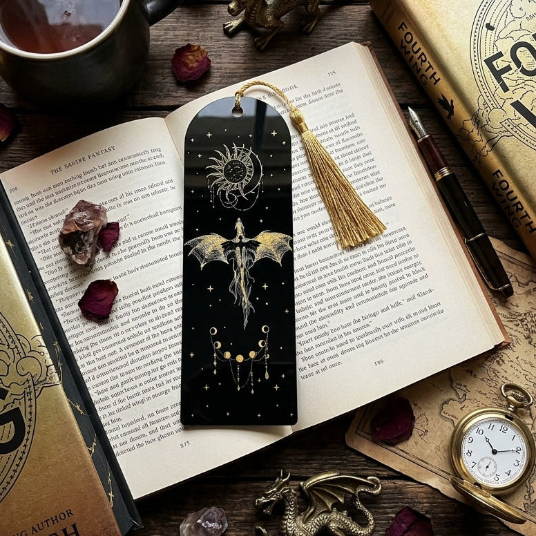 A black bookmark with a gold tassel and dragon illustration rests on an open book, surrounded by books, a cup of tea, crystals, dried roses, and fantasy-themed props on a wooden surface.