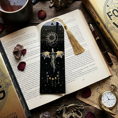 A black bookmark with a gold tassel and dragon illustration rests on an open book, surrounded by books, a cup of tea, crystals, dried roses, and fantasy-themed props on a wooden surface.