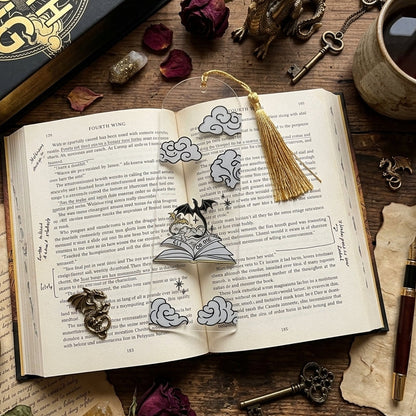 An open vintage-style book with a clear acrylic bookmark featuring an illustration of a dragons and phrase 'RIDE OR DIE', styled on a rustic wooden table with candles, keys, dried roses, and a cup of tea. A black book is visible in the top left.
