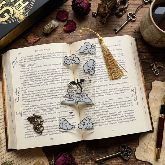 An open vintage-style book with a clear acrylic bookmark featuring an illustration of a dragons and phrase 'RIDE OR DIE', styled on a rustic wooden table with candles, keys, dried roses, and a cup of tea. A black book is visible in the top left.