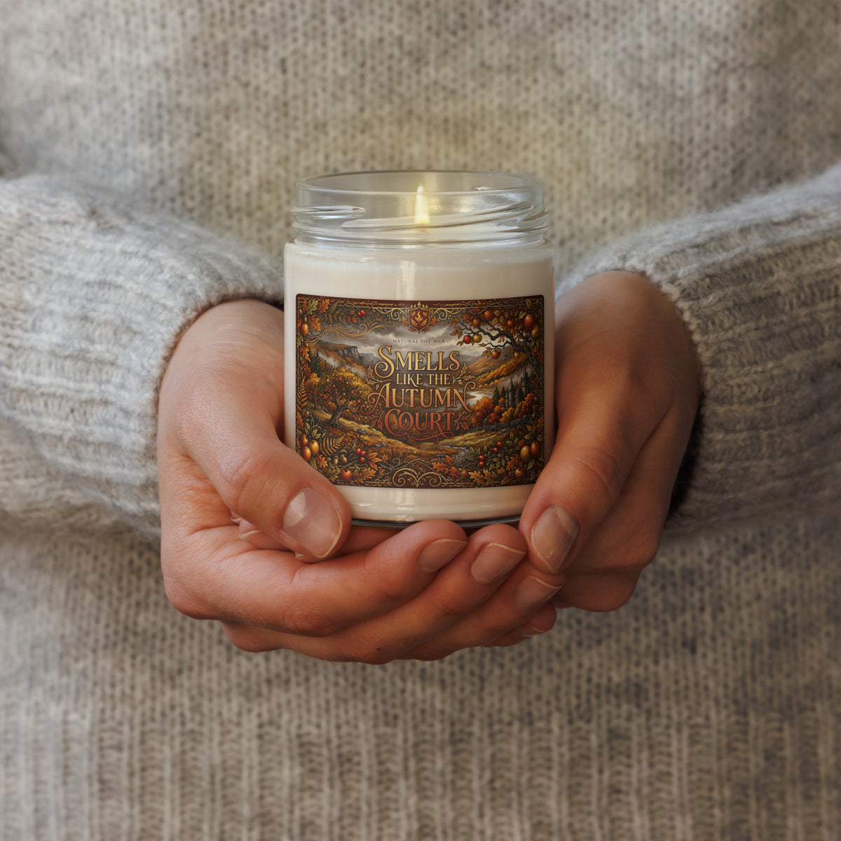 A person in a gray knit sweater holds a lit white candle in a clear glass jar. The label features a detailed autumn landscape illustration with the text 'SMELLS LIKE THE AUTUMN COURT'.