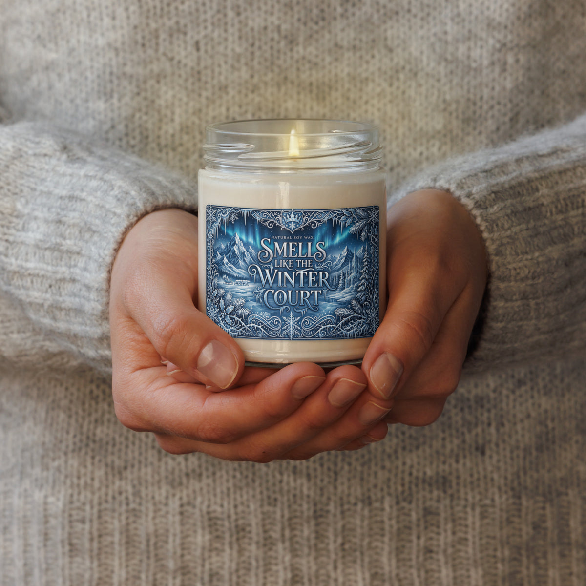 A person in a gray knit sweater holds a glass jar candle with a blue and white label that reads 'SMELLS LIKE THE WINTER COURT.'.