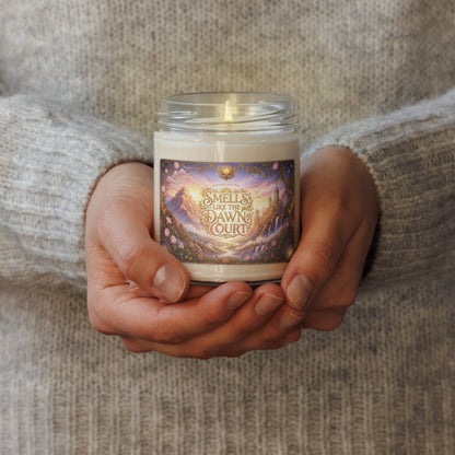 A person in a cozy gray knit sweater holds a lit glass jar candle. The candle has a colorful label reading 'Smells like the Dawn Court' with a fantasy landscape illustration in purple, gold, and blue tones.