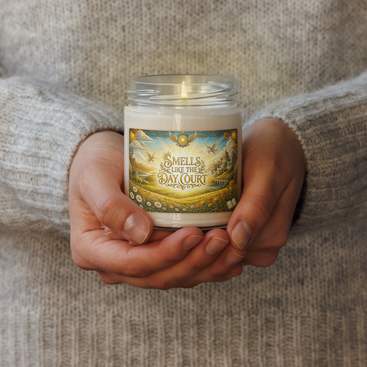 A person in a gray knit sweater holds a lit white candle in a clear glass jar. The front label reads 'SMELLS LIKE THE DAY COURT' with an illustrated landscape scene.