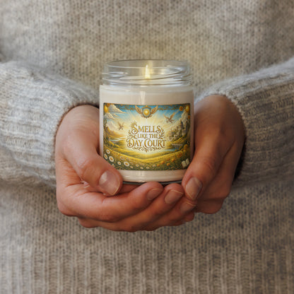 A person in a gray knit sweater holds a lit white candle in a clear glass jar. The front label reads 'SMELLS LIKE THE DAY COURT' with an illustrated landscape scene.