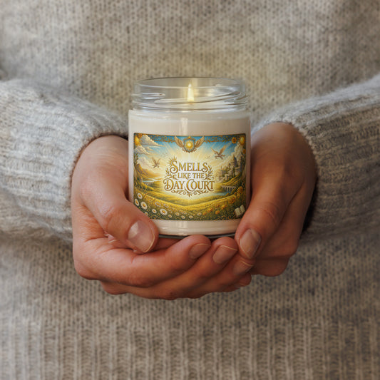 A person in a gray knit sweater holds a lit white candle in a clear glass jar. The front label reads 'SMELLS LIKE THE DAY COURT' with an illustrated landscape scene.