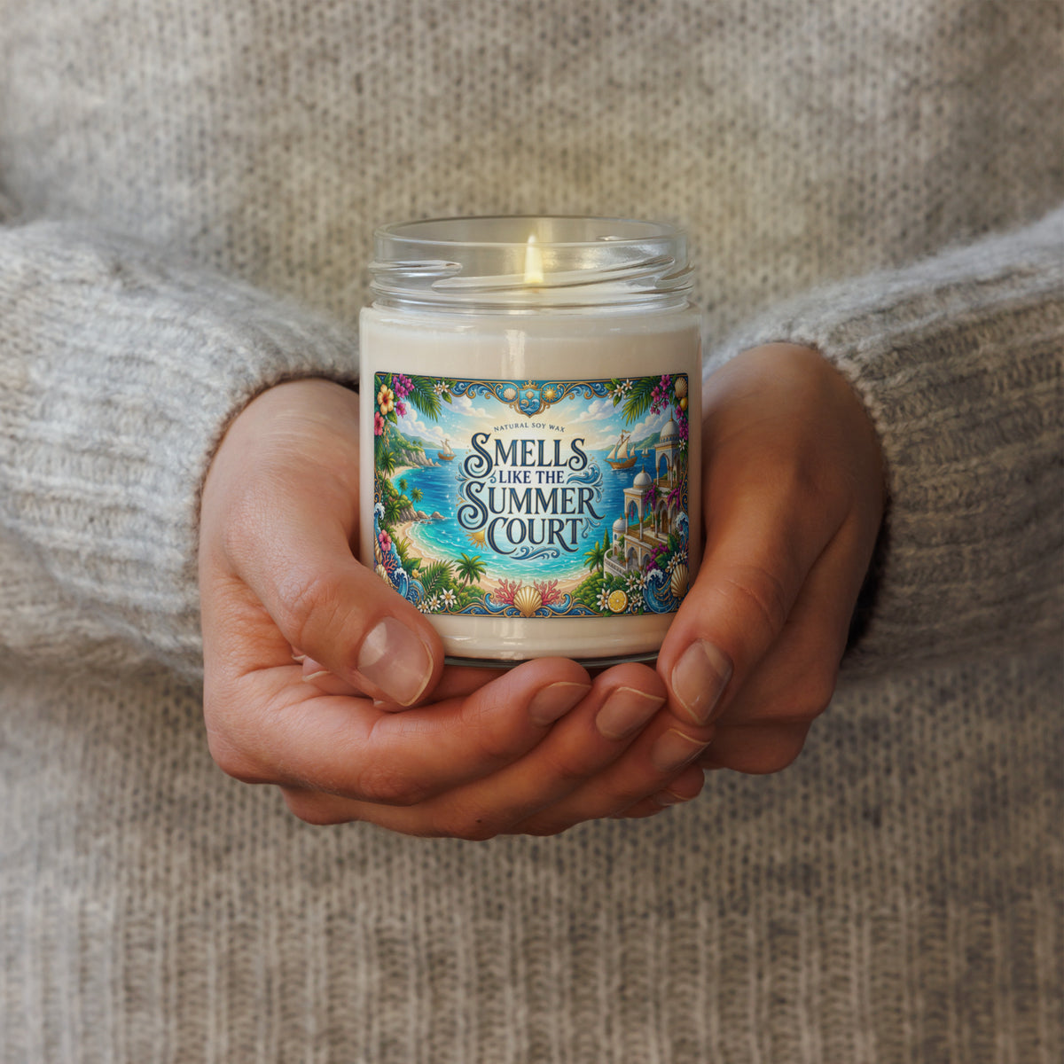 A person in a gray knit sweater holds a glass jar candle with a colorful illustrated label that reads “SMELLS LIKE THE SUMMER COURT.”.