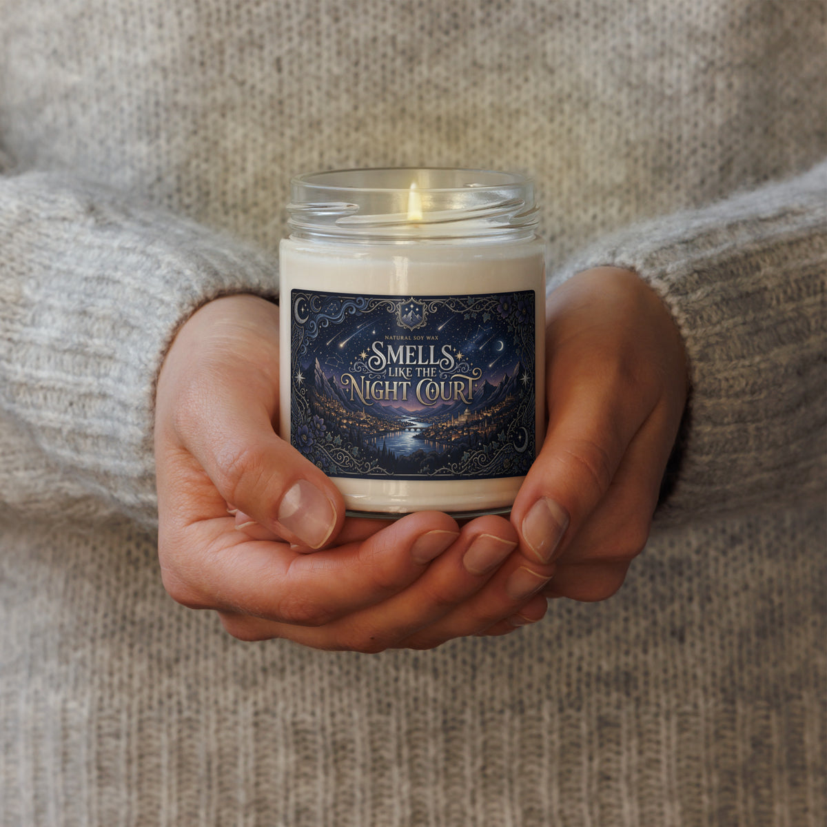 A person in a cozy gray knit sweater holds a lit candle in a clear glass jar. The candle has a cream-colored wax and a dark navy blue label with gold and white text that reads 'SMELLS LIKE THE NIGHT COURT'.