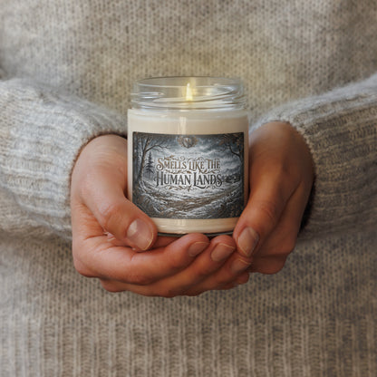 A person in a gray knit sweater holds a lit candle in a clear glass jar. The candle has an off-white wax body and a dark gray label with fantasy-style text reading 'SMELLS LIKE THE HUMAN LANDS.'