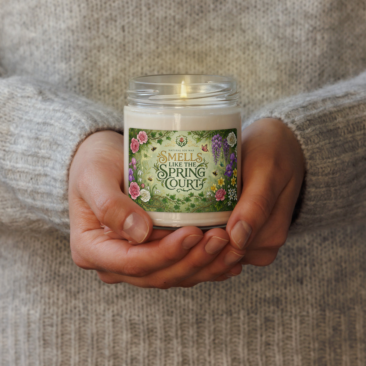 A person in a gray knit sweater holds a lit scented candle in a clear glass jar. The candle has a white wax body and a decorative green label with floral illustrations and the text 'SMELLS LIKE THE SPRING COURT.