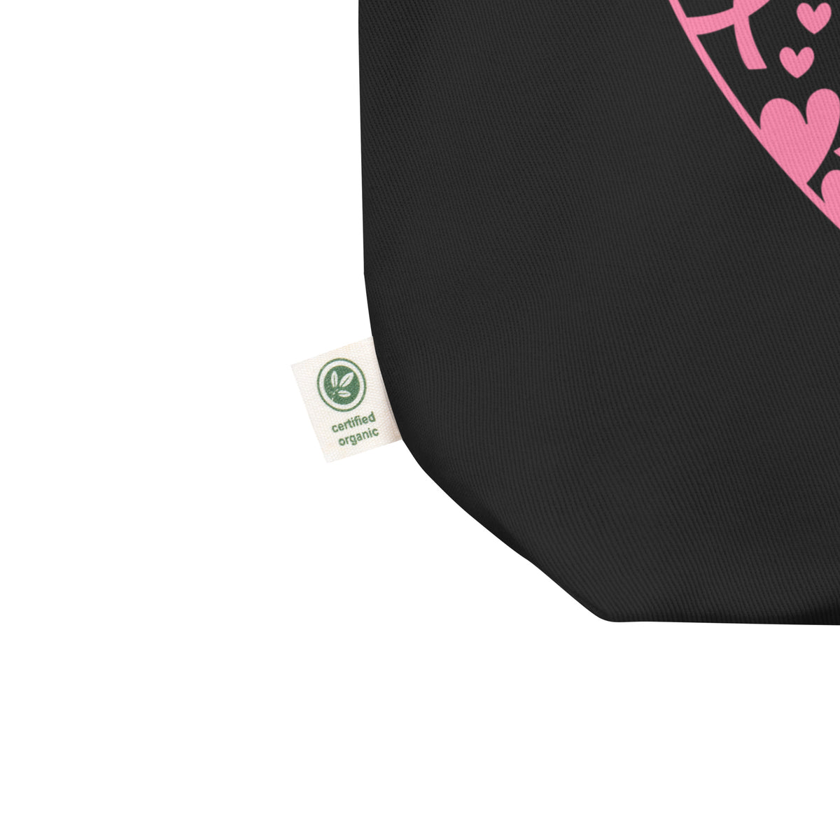 Close-up of a black fabric tote bag corner with a small white 'certified organic' tag and part of a pink heart graphic on a white background.