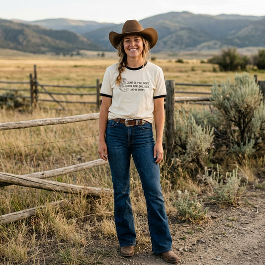 A woman stands outdoors in a rural landscape wearing a cream ringer T-shirt with black trim and a western-style graphic print that says, 'Some of ya'll don't know how Earl died, and it shows.', blue jeans, a brown cowboy hat, and brown boots. The background shows dry grass, a wooden fence, shrubs, and distant mountains.
