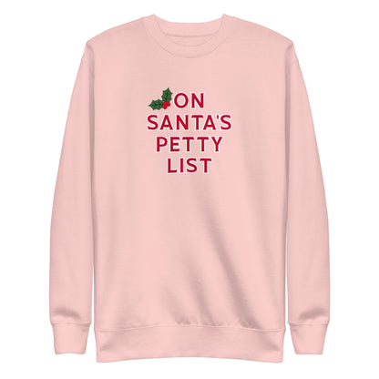 A light pink crewneck sweatshirt featuring the text 'ON SANTA'S PETTY LIST' in red, with a small green holly graphic at the top left of the text, shown on a plain white background.