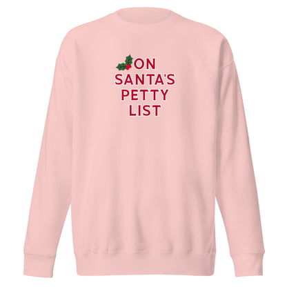 A light pink crewneck sweatshirt featuring the text 'ON SANTA'S PETTY LIST' in red, with a small green and red holly graphic above the text, displayed on a solid white background.