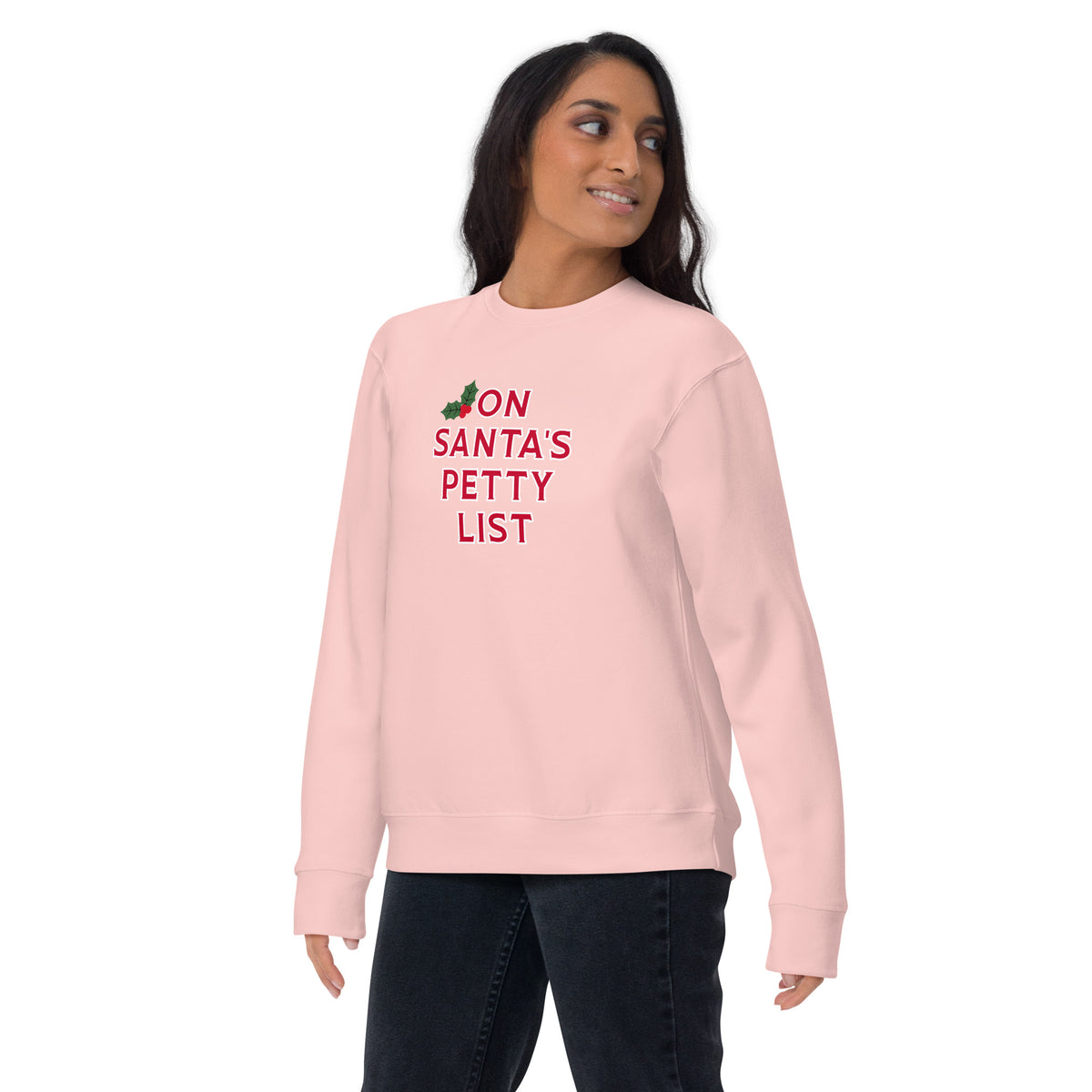 A woman modeling a light pink crewneck sweatshirt with the text 'ON SANTA'S PETTY LIST' printed in red on the chest, paired with dark jeans against a plain white background.