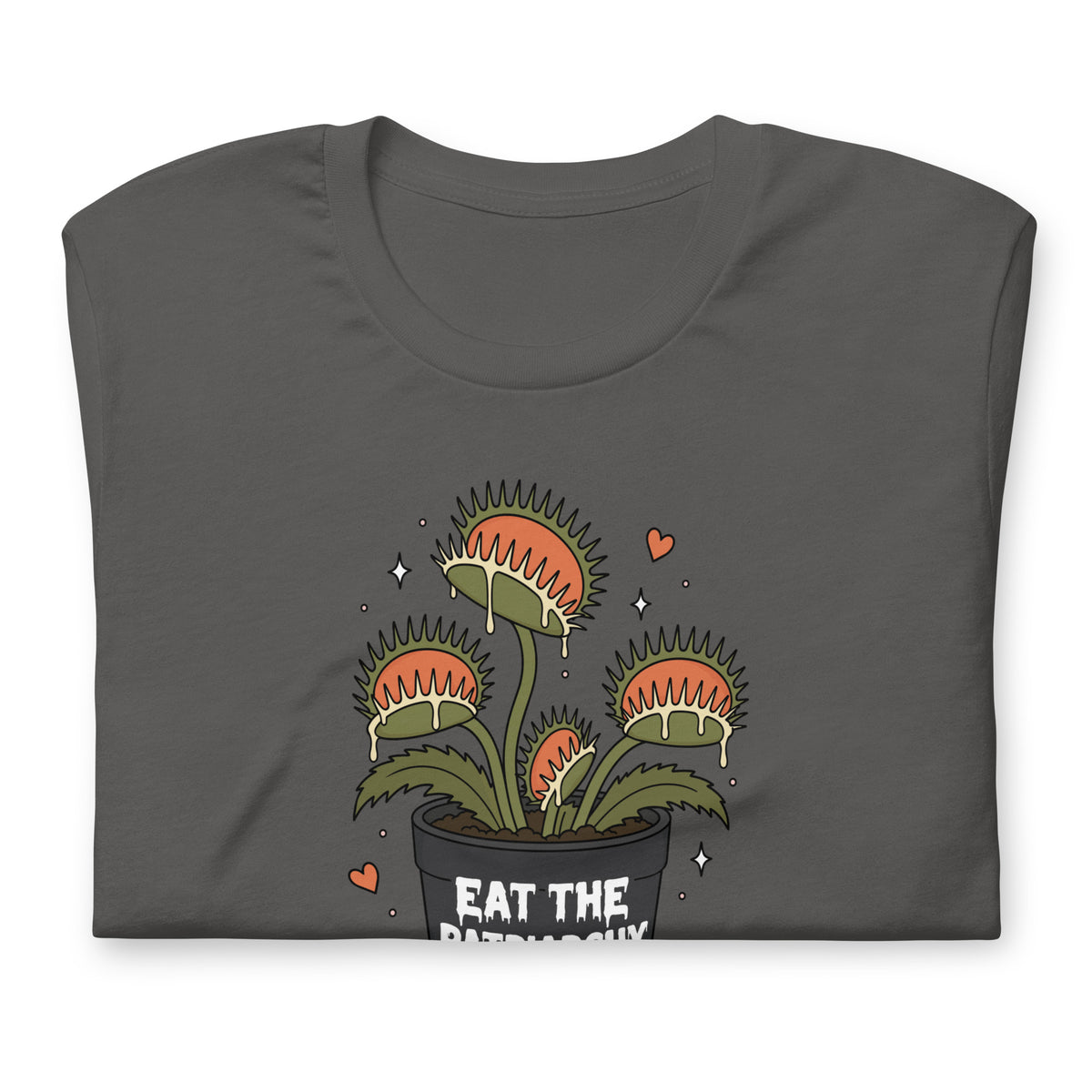 A dark gray folded t-shirt featuring a cartoon illustration of three Venus flytraps in a black pot with the text 'EAT THE PATRIARCHY' and small pink hearts and white stars.