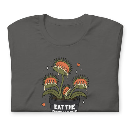 A dark gray folded t-shirt featuring a cartoon illustration of three Venus flytraps in a black pot with the text 'EAT THE PATRIARCHY' and small pink hearts and white stars.