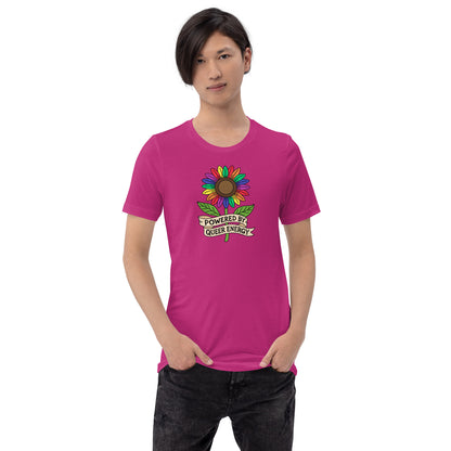 A man wearing a bright pink t-shirt with a colorful sunflower graphic and the text 'POWERED BY QUEER ENERGY' on the chest, paired with black jeans, against a plain white background.