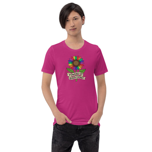 A man wearing a bright pink t-shirt with a colorful sunflower graphic and the text 'POWERED BY QUEER ENERGY' on the chest, paired with black jeans, against a plain white background.