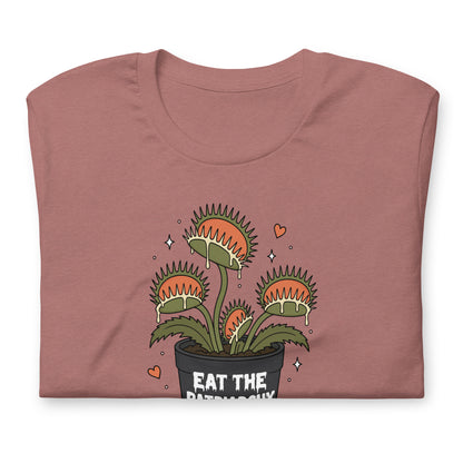 A folded dusty rose t-shirt with a cartoon graphic of a Venus flytrap plant in a black pot with the text 'EAT THE PATRIARCHY' and small orange hearts, shown on a plain white background.